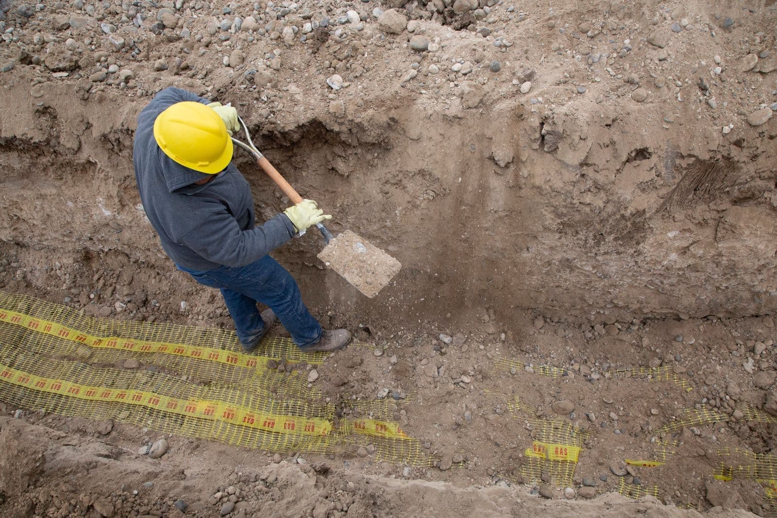 Waterproofing company crew member in a hard hat shovelling at the bottom of a deep trench